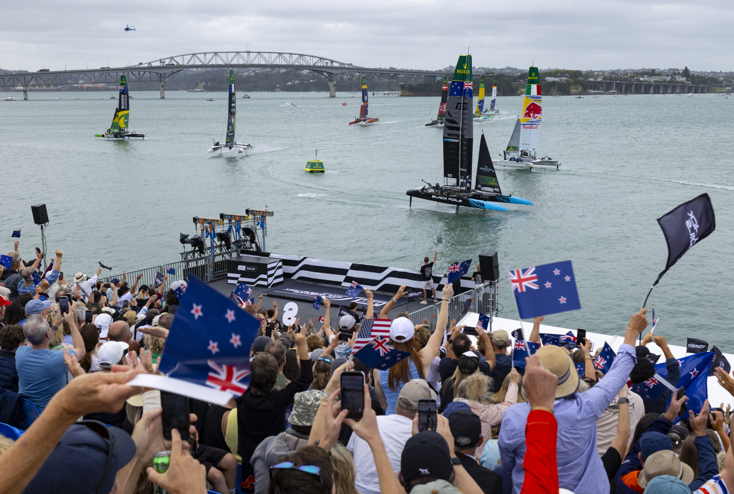 Spectators watch from the grandstand as the fleet led by Black Foils SailGP Team helmed by Peter Burling race past on Race Day 1 of the ITM New Zealand Sail Grand Prix in Auckland, New Zealand. Saturday 14 February 2026. Rolex SailGP Championship Event 2 2026 Season. Photo: Brett Phibbs for SailGP. Handout image supplied by SailGP